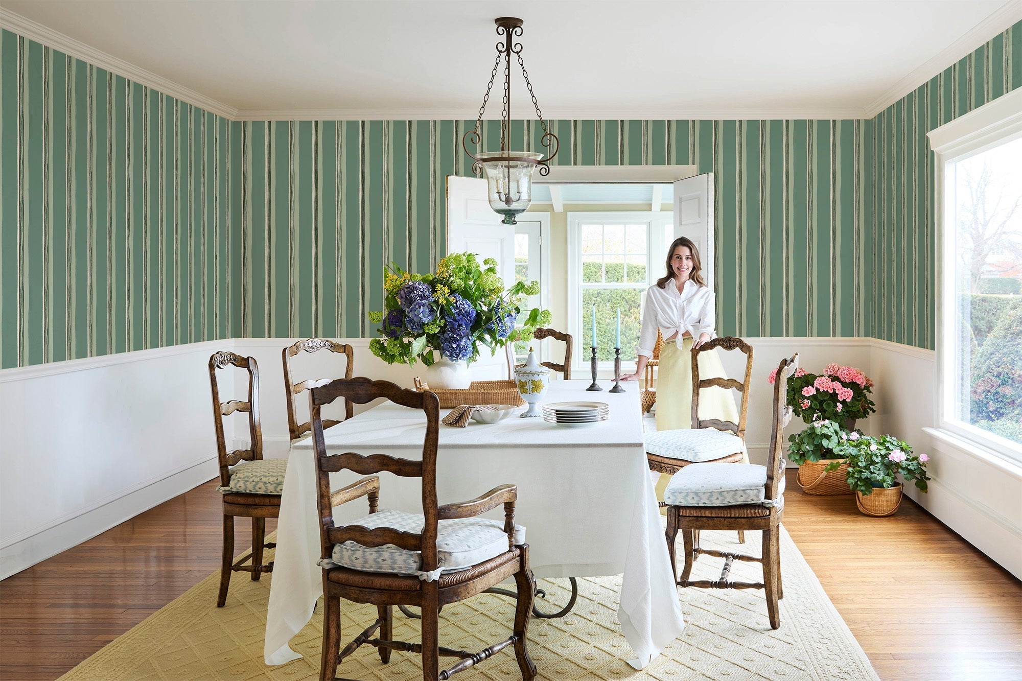 Salle à manger lumineuse aux murs à rayures vertes et blanches, grande table nappée de blanc entourée de chaises en bois ancien, bouquet de fleurs au centre, suspension au plafond et femme souriante debout près de la table.